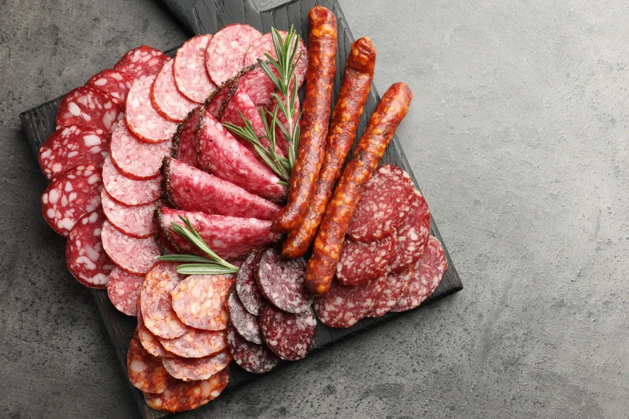Tábua de charcutaria com carnes fatiadas e linguiças, decorada com raminhos de alecrim, sobre uma superfície cinza escura.