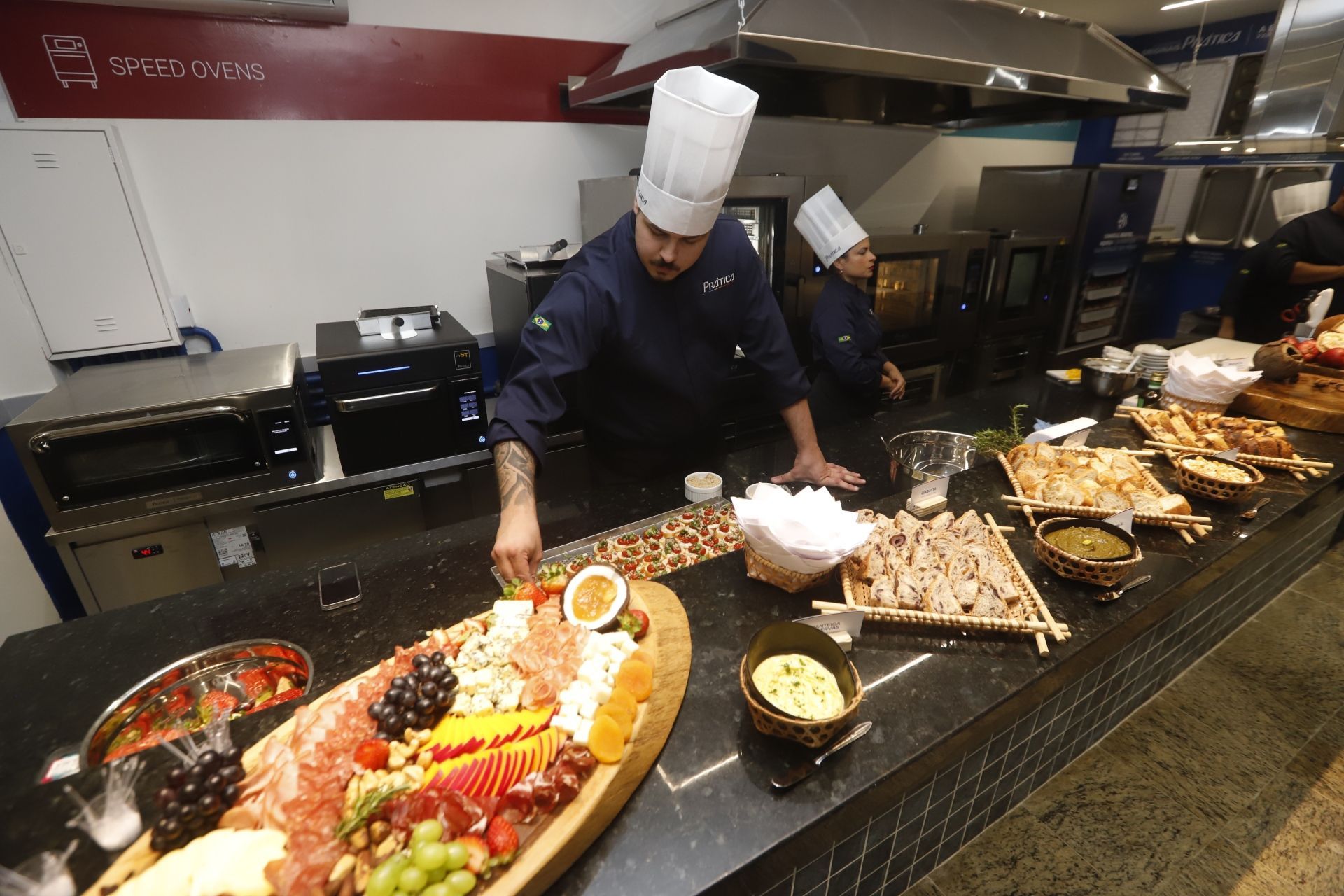 Chefs preparando comida em um balcão. Um arruma uma tábua de frios, o outro prepara a comida em uma cozinha comercial.