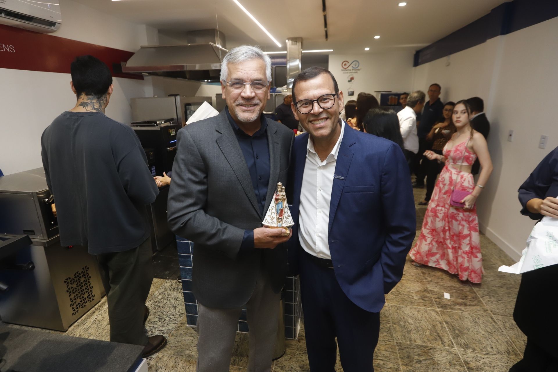 Dois homens posam em ambiente fechado, um deles segurando um troféu. Outras pessoas ao fundo.