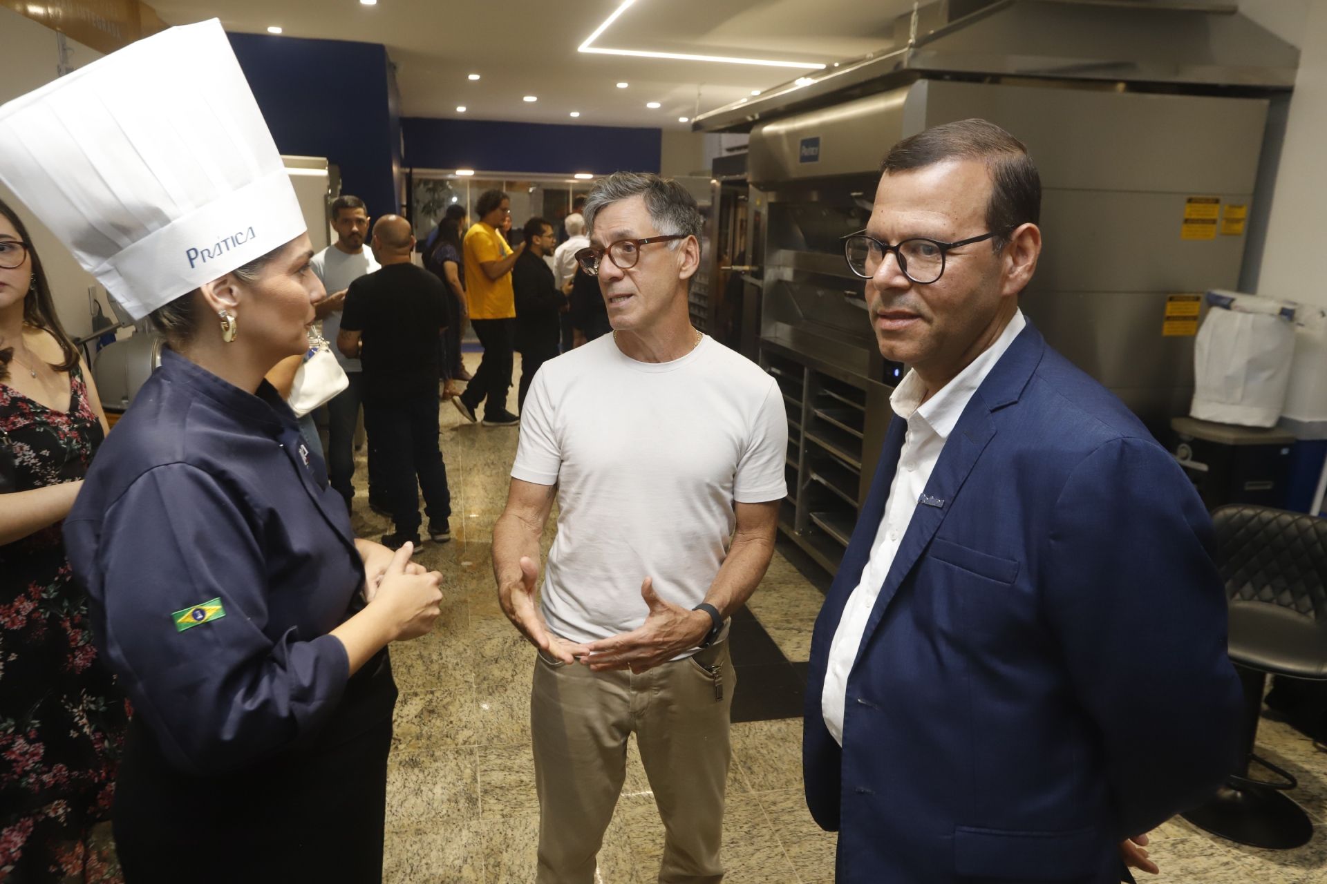 Chef uniformizado conversando com dois homens na cozinha, outras pessoas ao fundo.