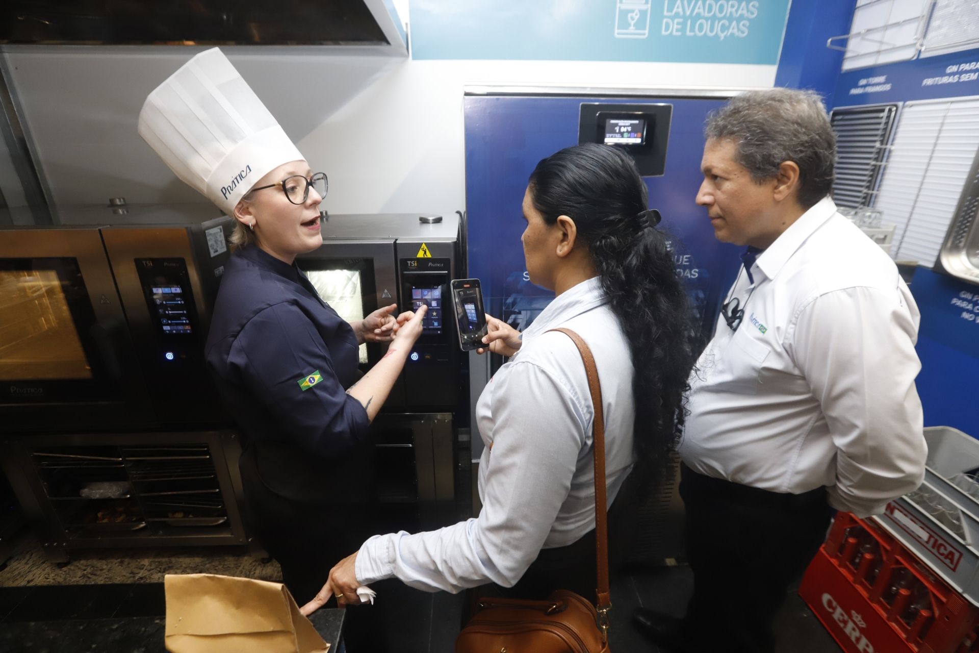 Um chef de branco explica um forno comercial para duas pessoas. Ambiente de cozinha, cores escuras.