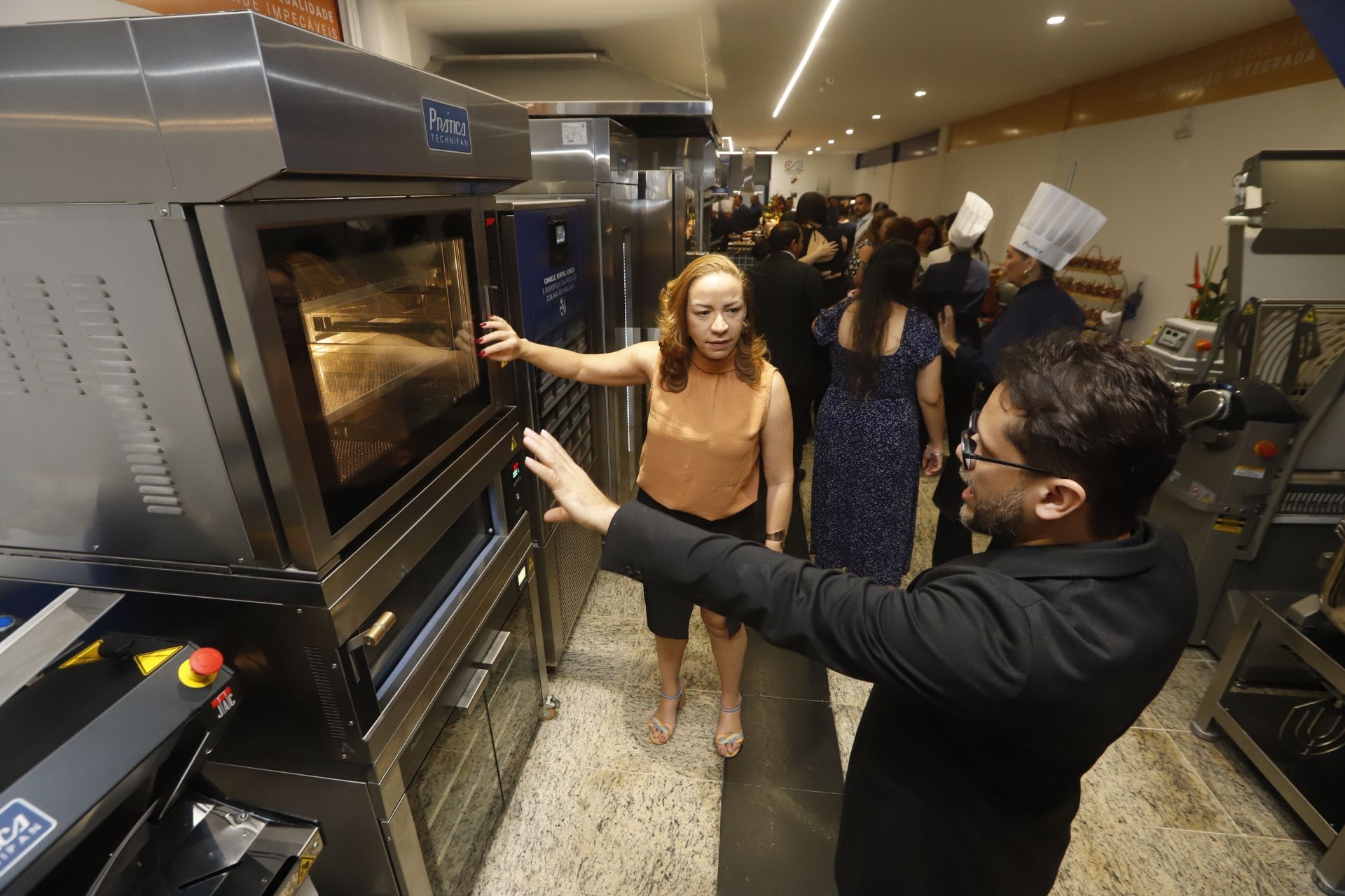 Mulher e homem examinando um forno em uma cozinha comercial. Outras pessoas e equipamentos são visíveis ao fundo.