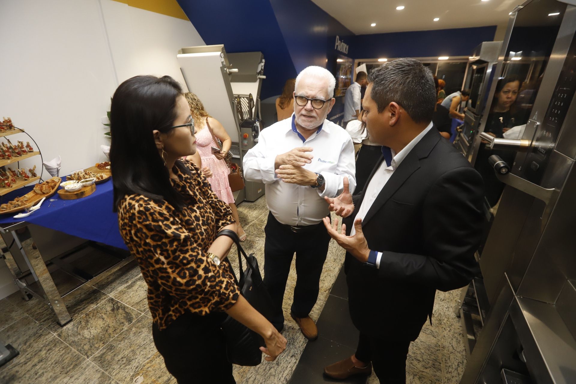 Três pessoas conversam em uma padaria: uma mulher com estampa de oncinha, um homem de camisa branca e um homem de terno.