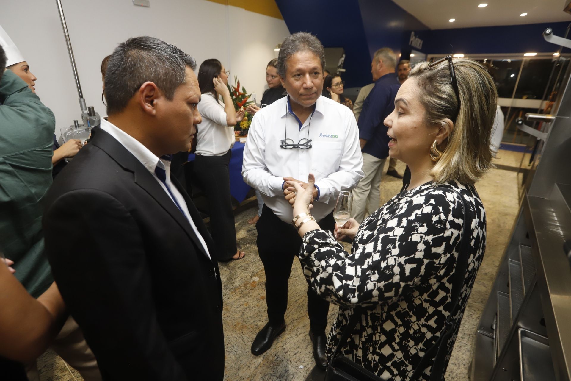 Pessoas em trajes executivos conversando em um evento. Espaço interno com detalhes em azul e amarelo.