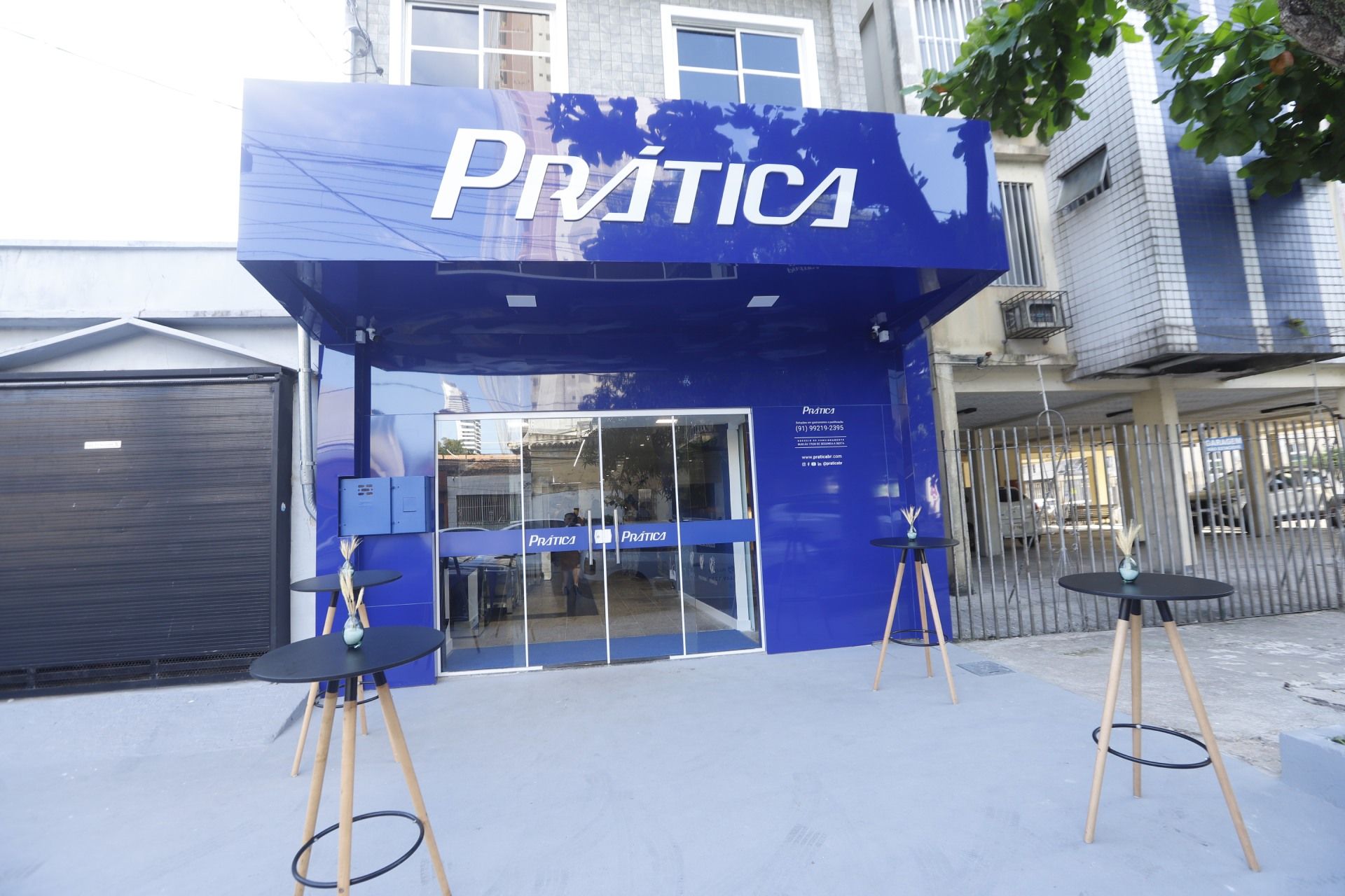 Fachada da loja Prática em azul e branco com mesas ao ar livre e porta de garagem fechada.