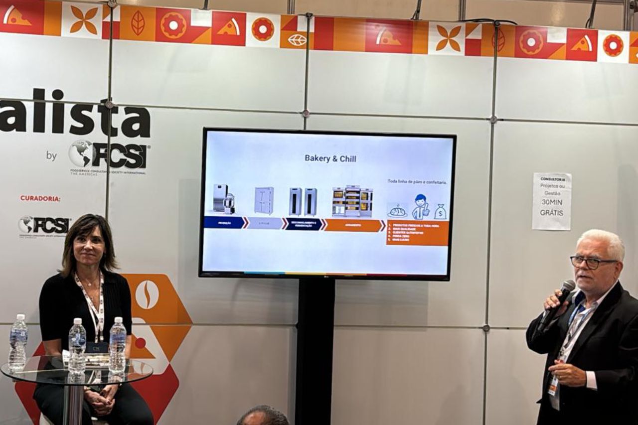 Um homem está falando em um microfone enquanto uma mulher está sentada atrás dele