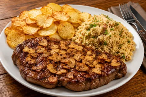 Bife grelhado com batatas fritas e arroz em um prato branco com talheres.