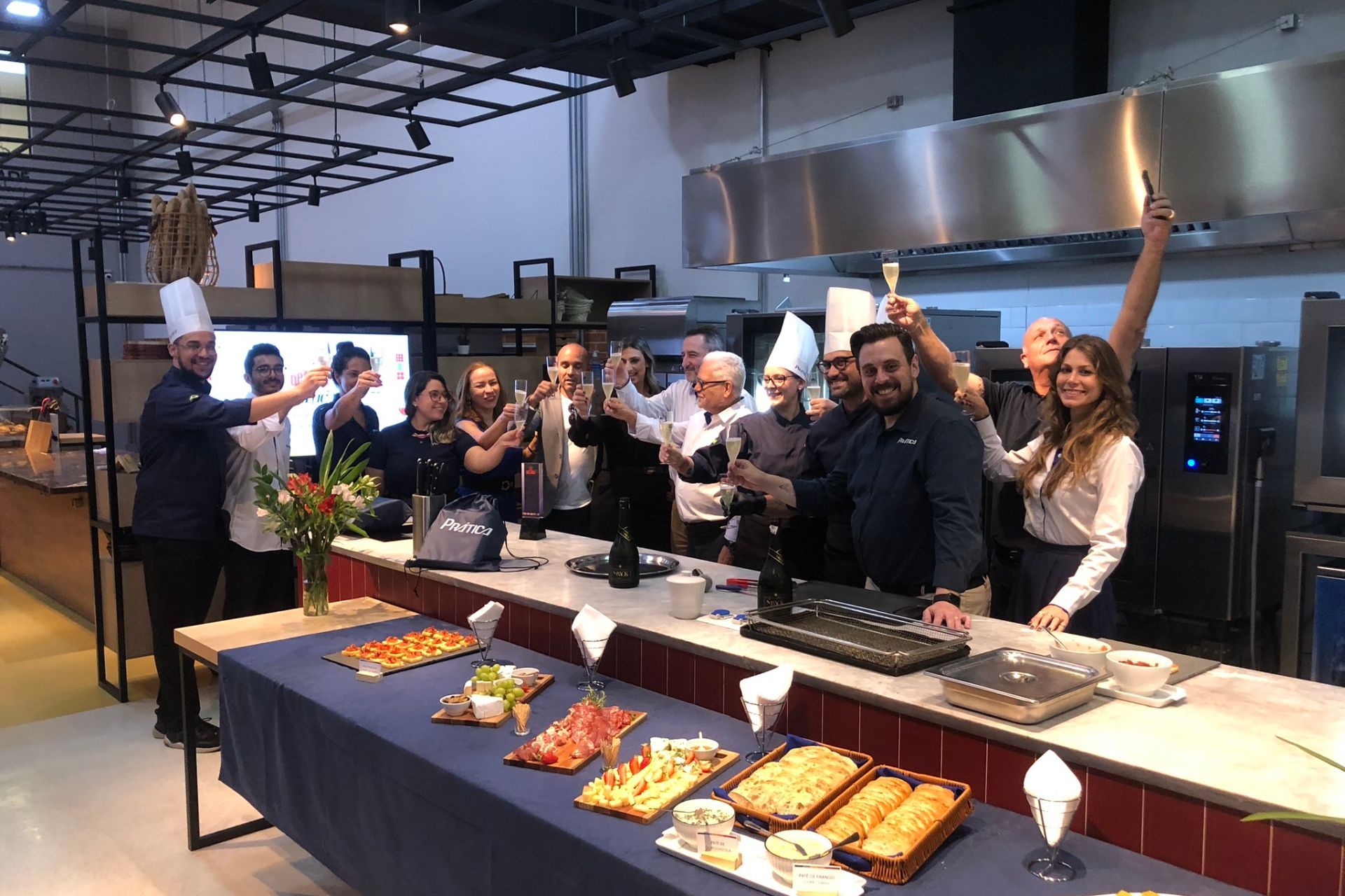 Grupo comemorando em uma cozinha. As pessoas brindam, algumas usam chapéus de chef. Há comida sobre a mesa.