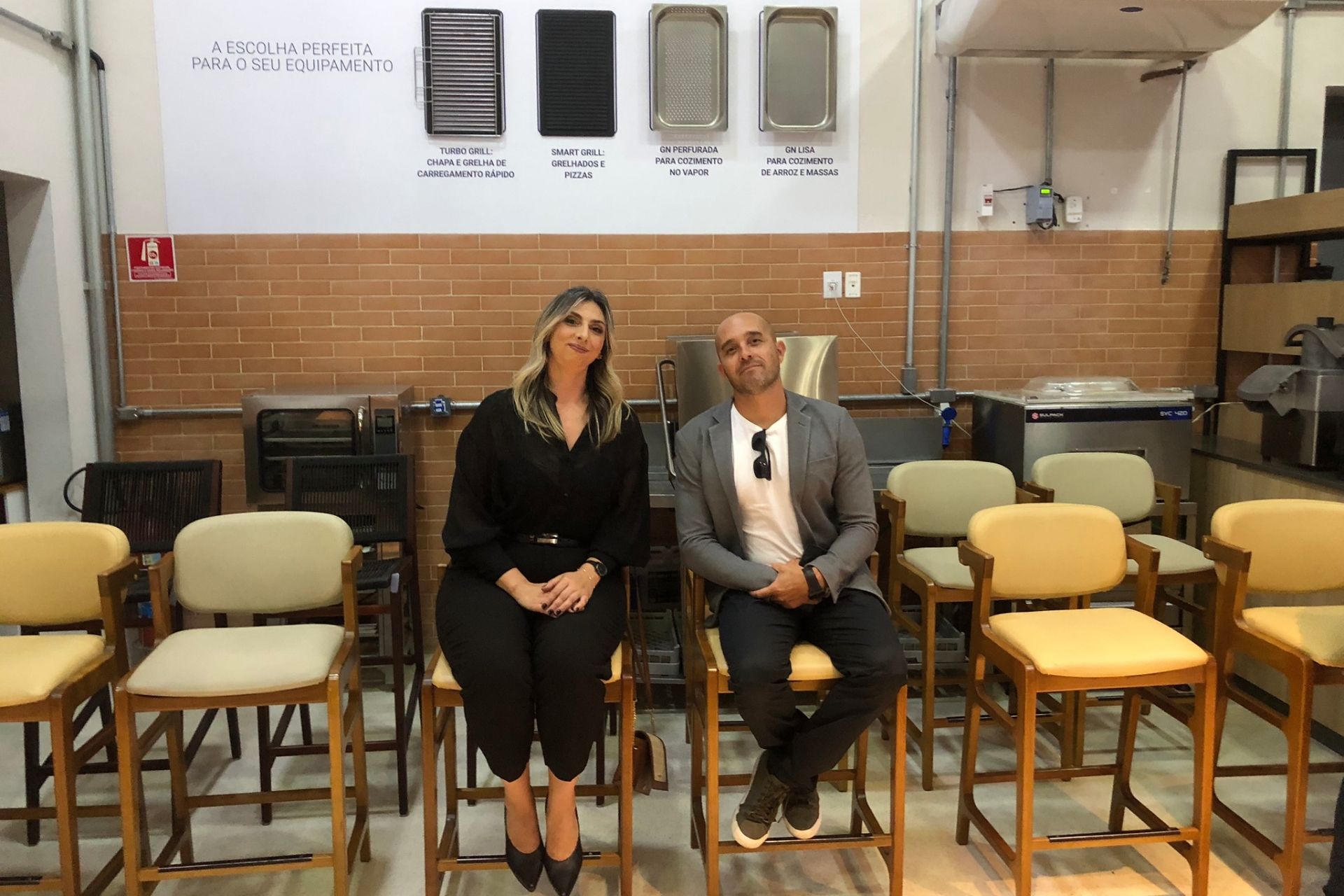 Duas pessoas sentadas em bancos em uma oficina, olhando para a câmera. Amostras estão expostas na parede atrás delas.