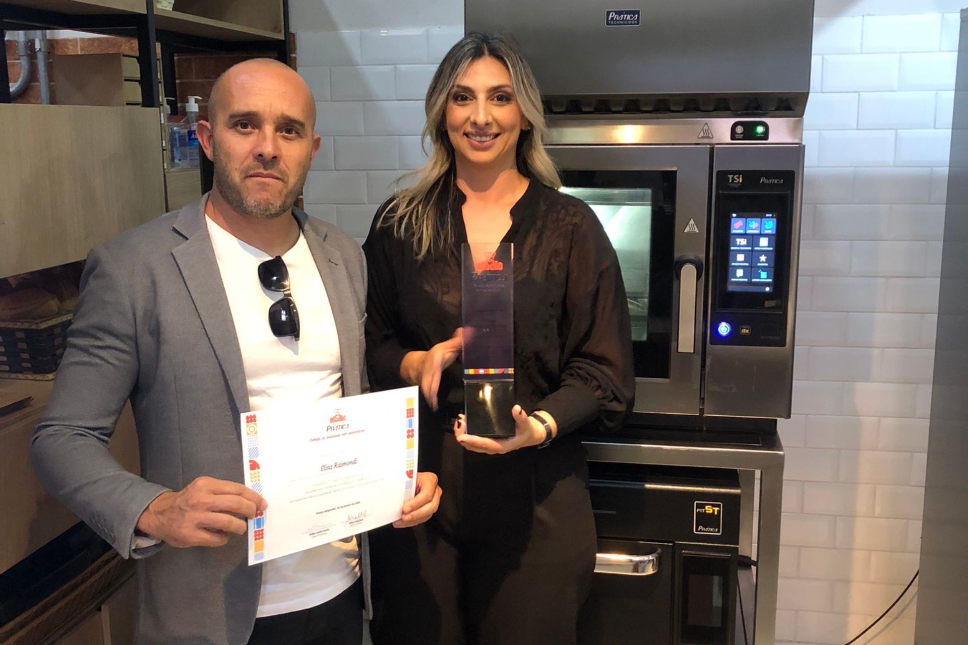 Homem e mulher segurando um prêmio e um certificado perto de um forno em uma cozinha.