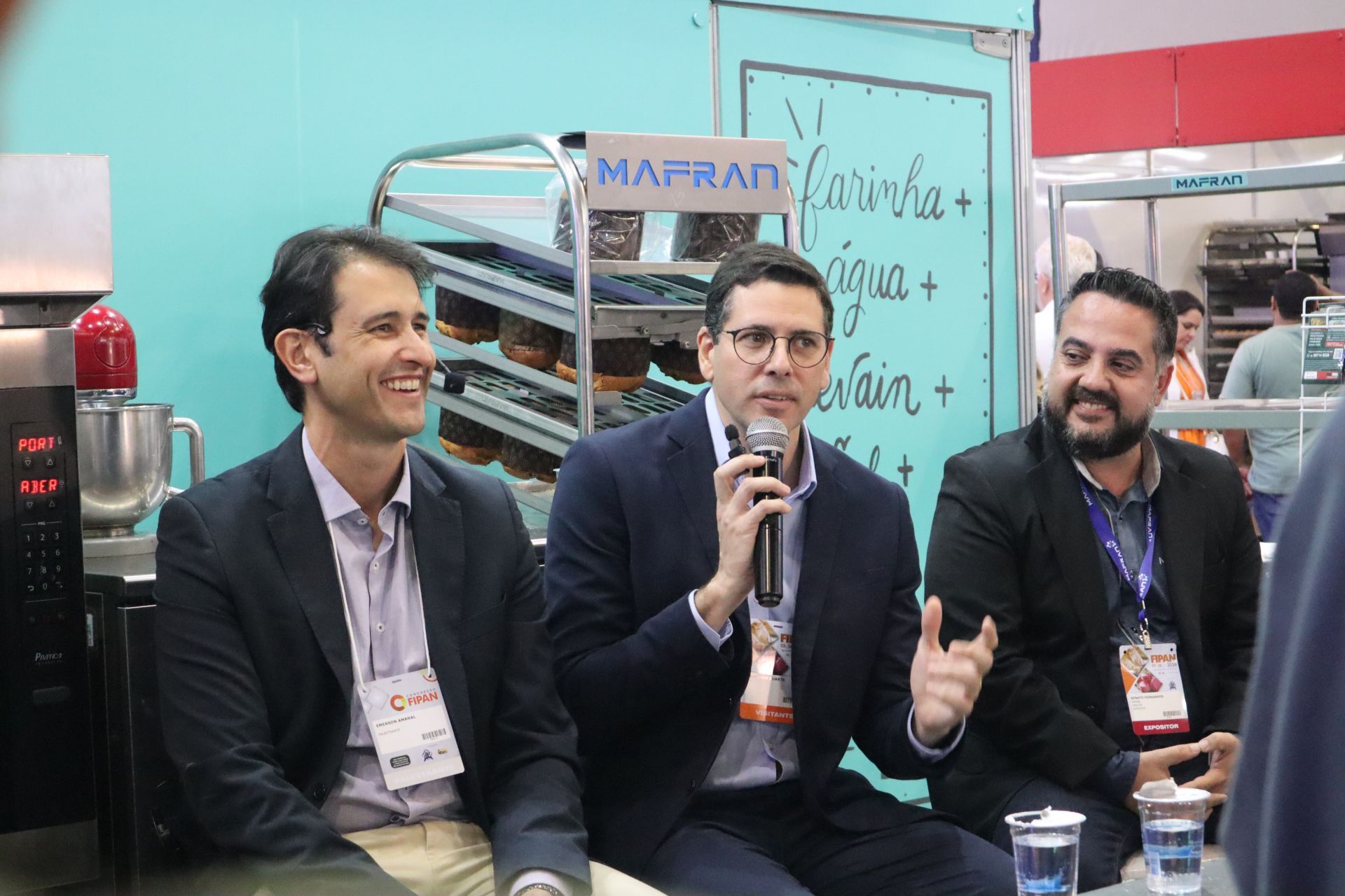 Três homens estão sentados à mesa falando em microfones.