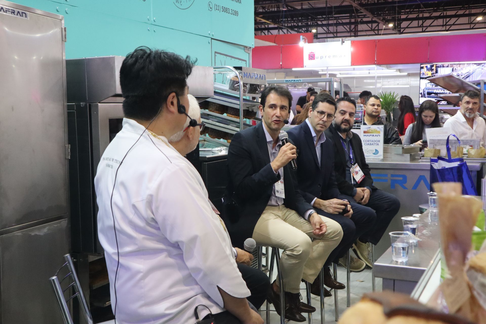 Um homem está falando em um microfone enquanto está sentado na frente de um grupo de pessoas.