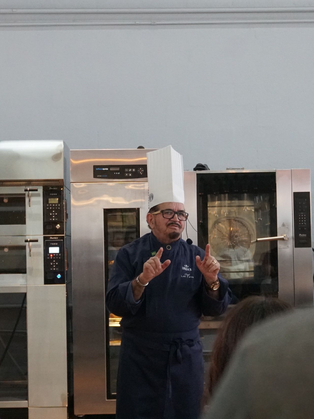 Chef de chapéu branco e uniforme azul falando e gesticulando em frente aos fornos.