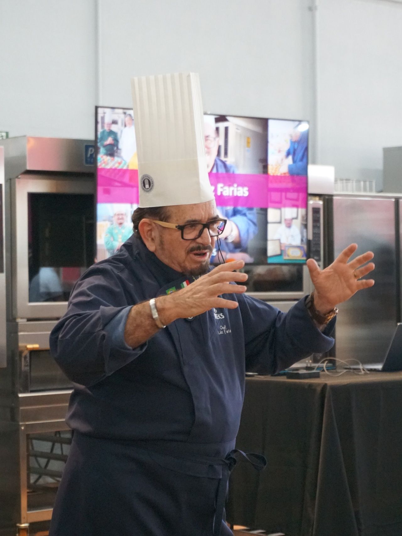 Chef falando, gesticulando com as mãos em uma cozinha. Usando chapéu branco alto, óculos e uniforme azul de chef.