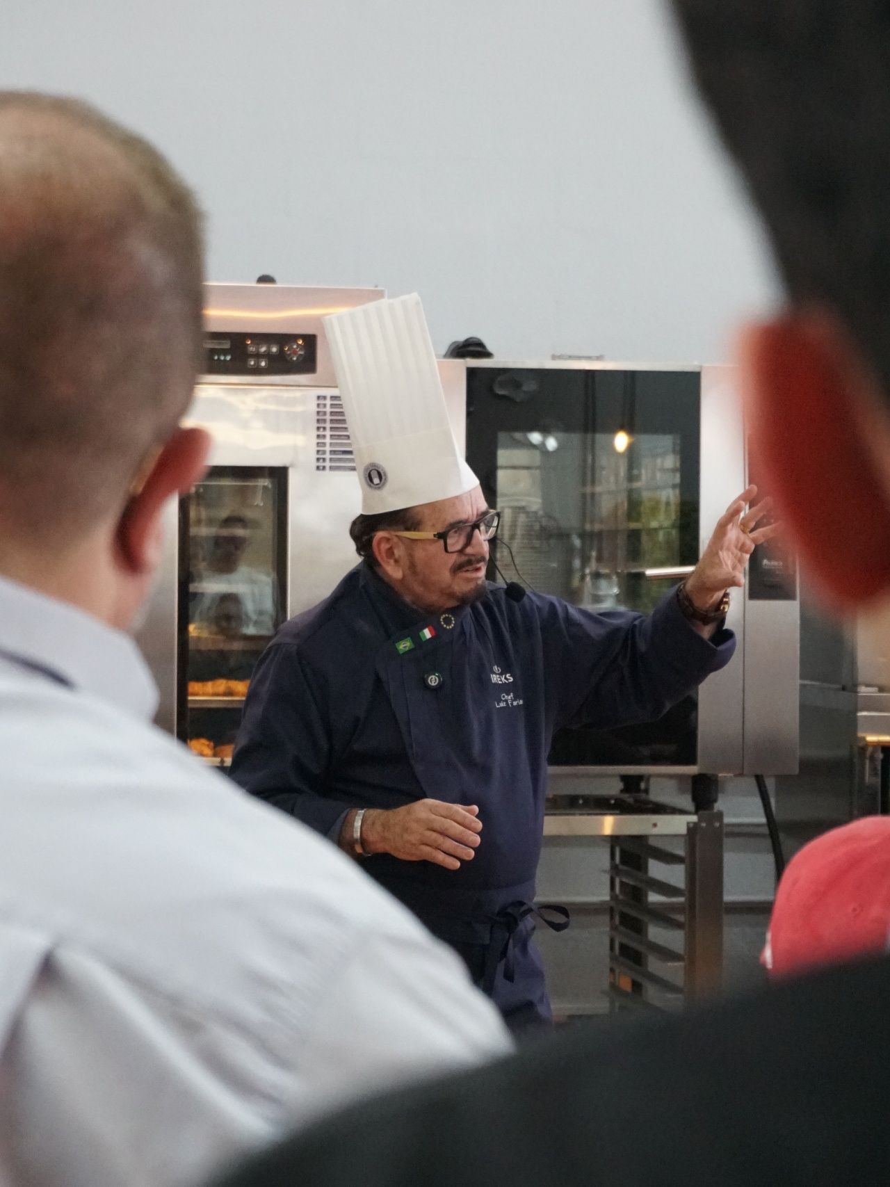 Chef de chapéu alto gesticula em direção aos fornos, falando para um grupo.