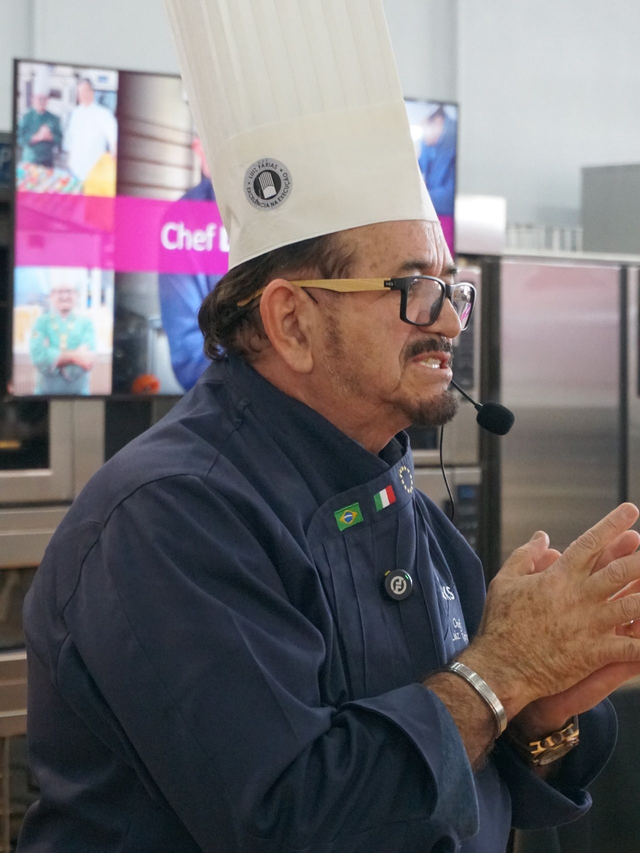 Chef, usando um chapéu branco alto e óculos, fala animadamente na cozinha.