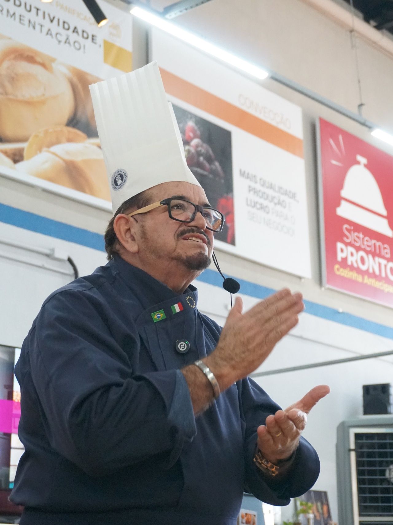 Chef de chapéu branco alto e óculos batendo palmas durante uma demonstração em uma padaria.