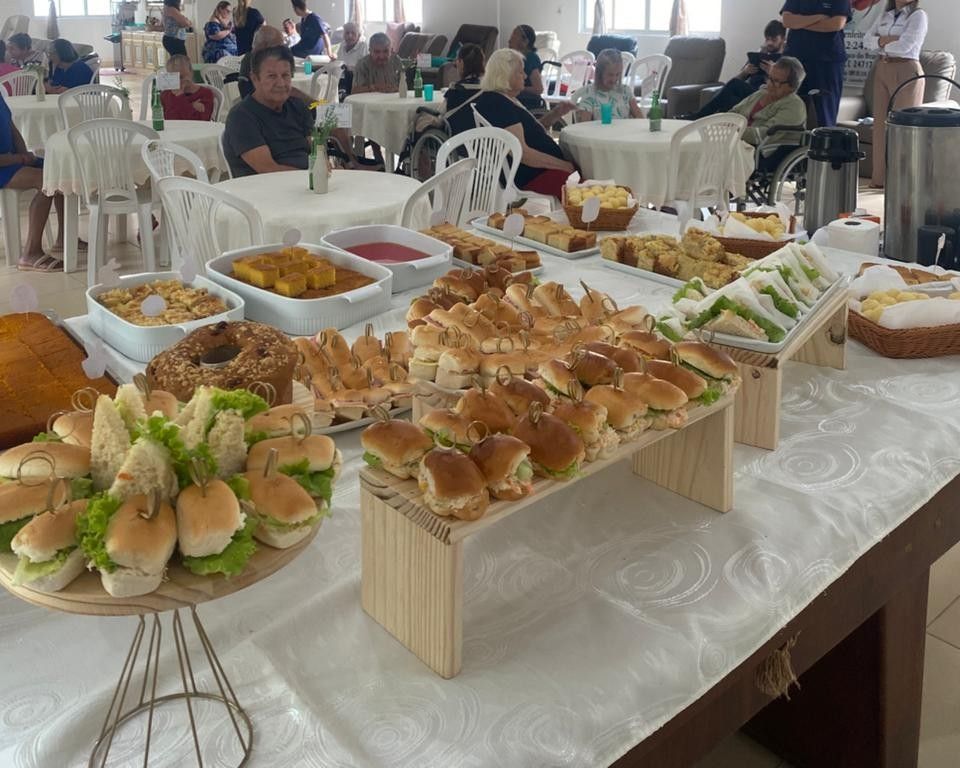 Uma mesa de bufê repleta de sanduíches, bolos e outros petiscos em uma reunião social em uma sala com convidados sentados.