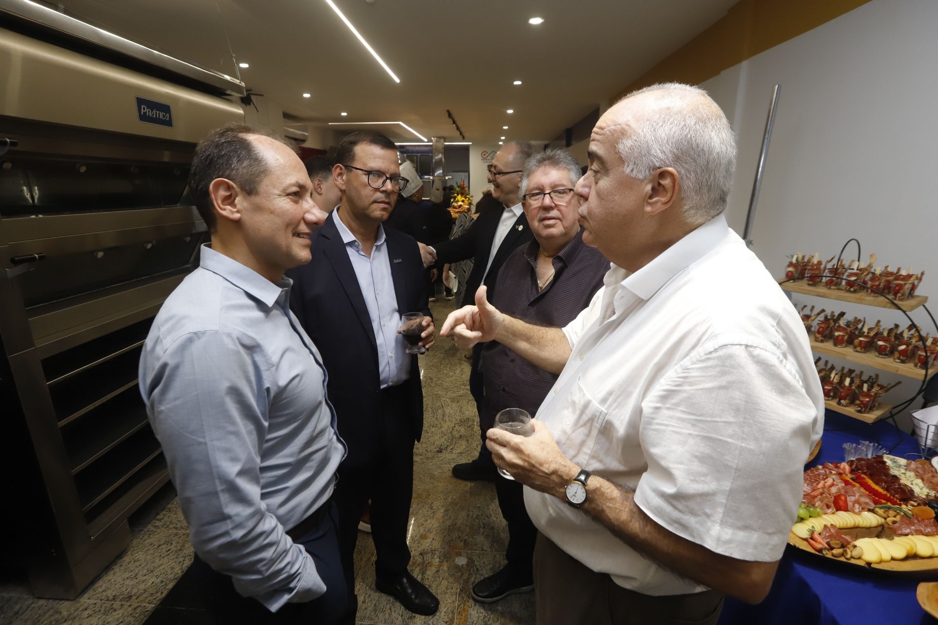 Homens em trajes formais conversando, possivelmente em um evento de networking. Pratos de queijo e carne são visíveis.