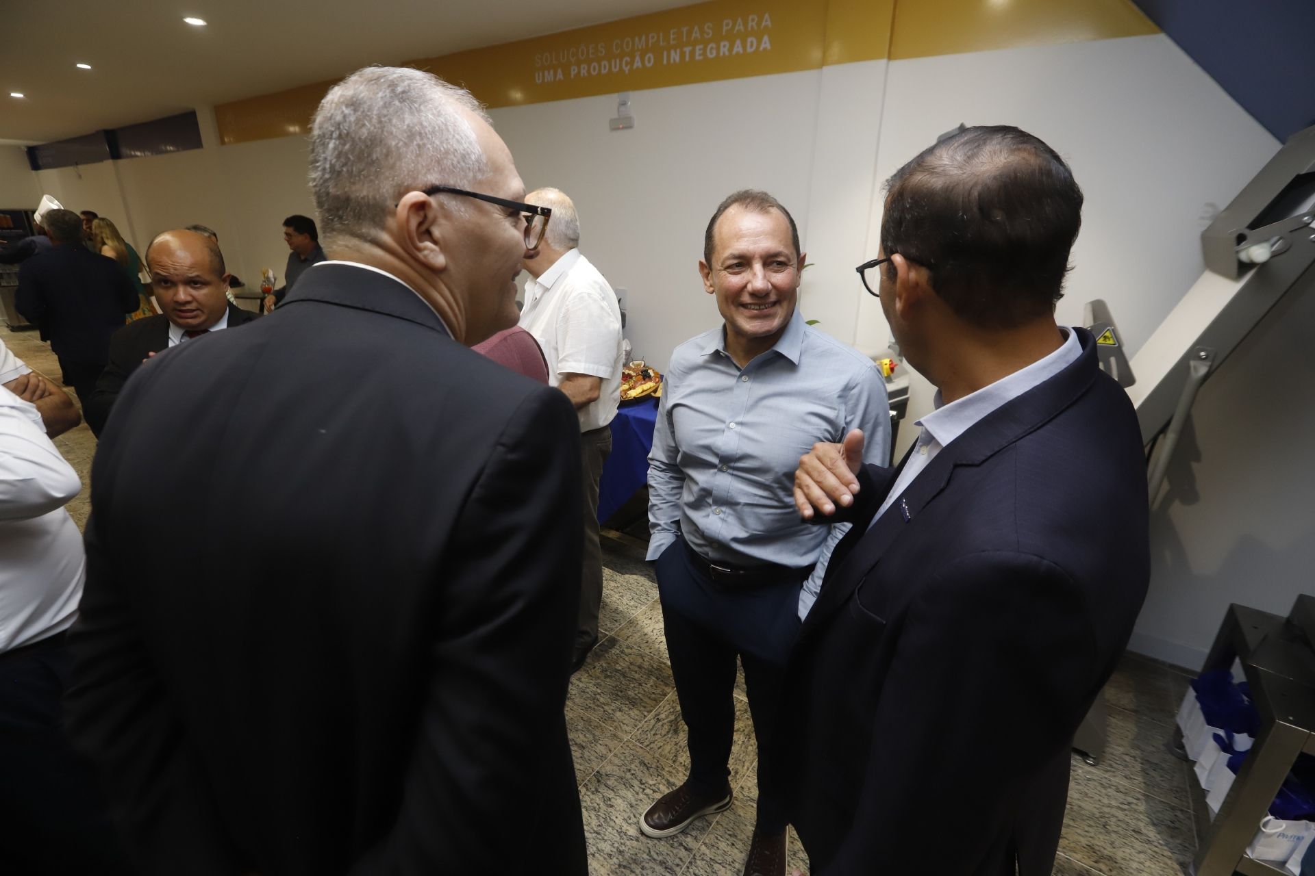 Três homens estão conversando em uma sala: um está de terno, um está de camisa social e outro de terno.