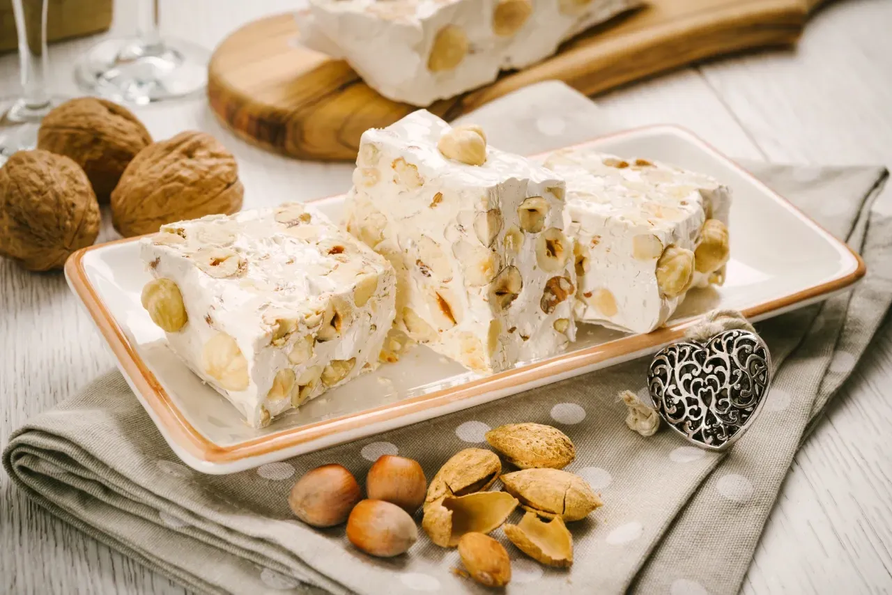 Nougat branco com nozes em um prato branco, rodeado por nozes e amêndoas.