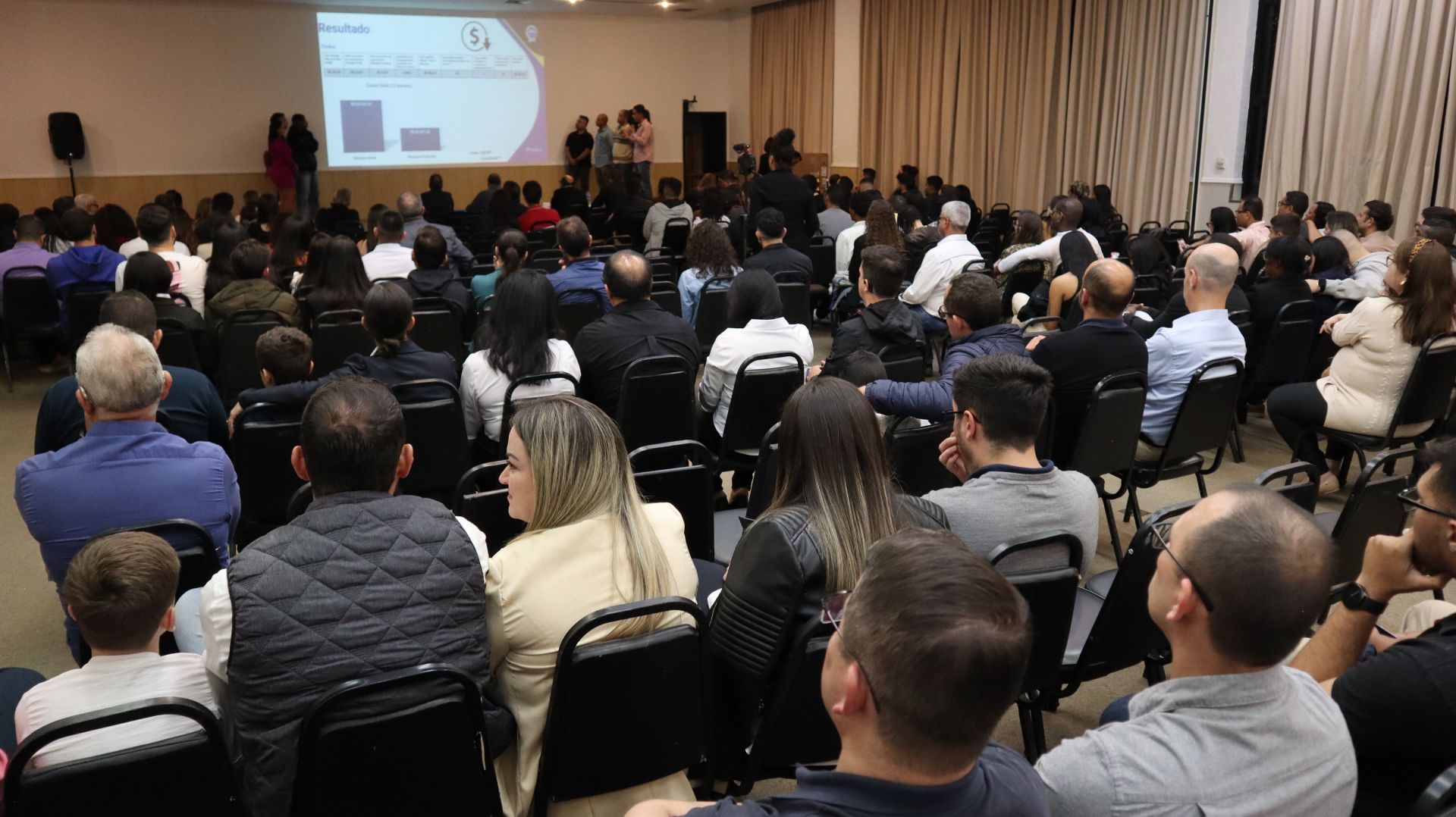 Um grande grupo de pessoas está sentado em uma sala de conferências assistindo a uma apresentação.