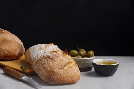 Pão crocante com azeitonas e azeite de oliva sobre uma tábua de madeira e mesa branca.