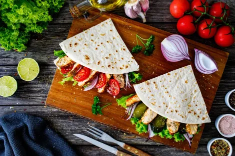 Duas piadinas dobradas, recheadas com frango grelhado e legumes, dispostas sobre uma tábua de madeira e rodeadas por ingredientes frescos.