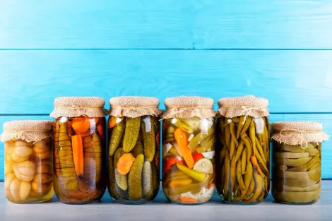 Seis potes de legumes em conserva variados sobre uma mesa contra um fundo de madeira azul.