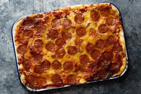 Pizza retangular de pepperoni servida em uma bandeja de metal, com queijo gratinado e crosta crocante.