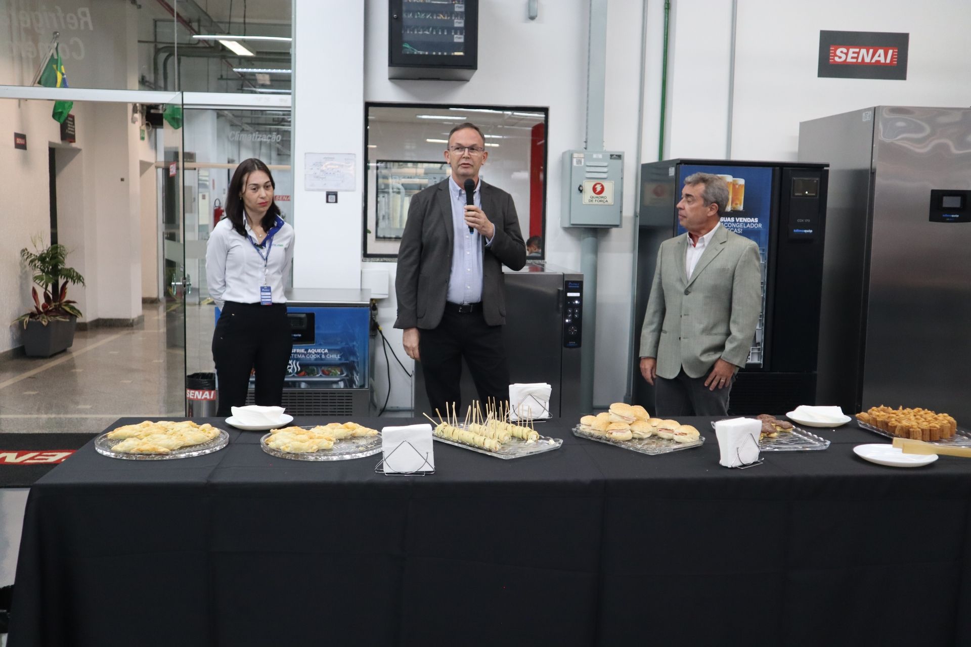 Três pessoas sentadas a uma mesa de recepção com comida. Um homem com um microfone fala, enquanto outros dois observam. Dentro de um prédio.