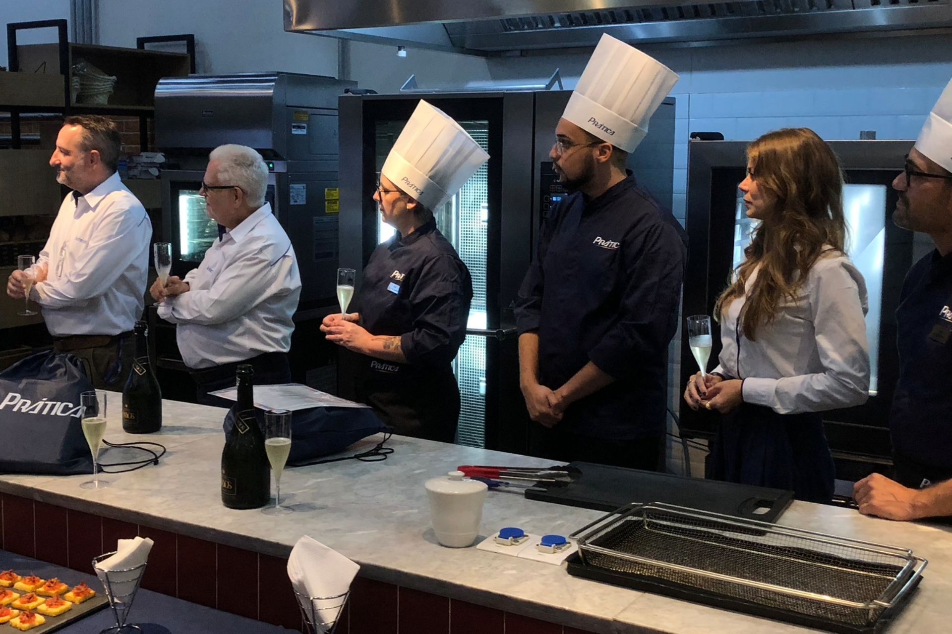Chefs e outros funcionários brindam com champanhe na cozinha. Pessoas estão atrás de um balcão com comida.