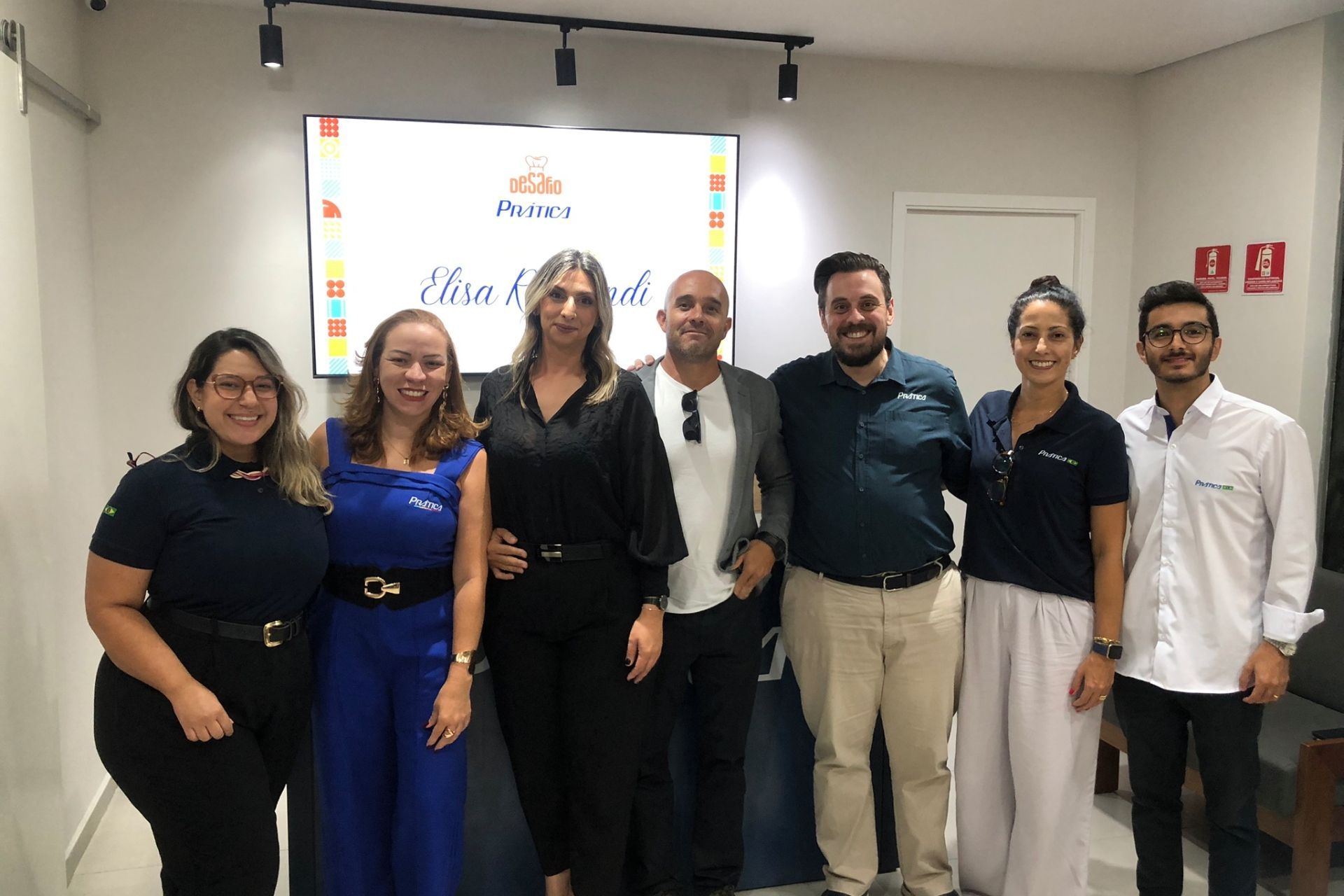Grupo de pessoas posando em frente a uma placa com um logotipo e texto, provavelmente em um ambiente de escritório.