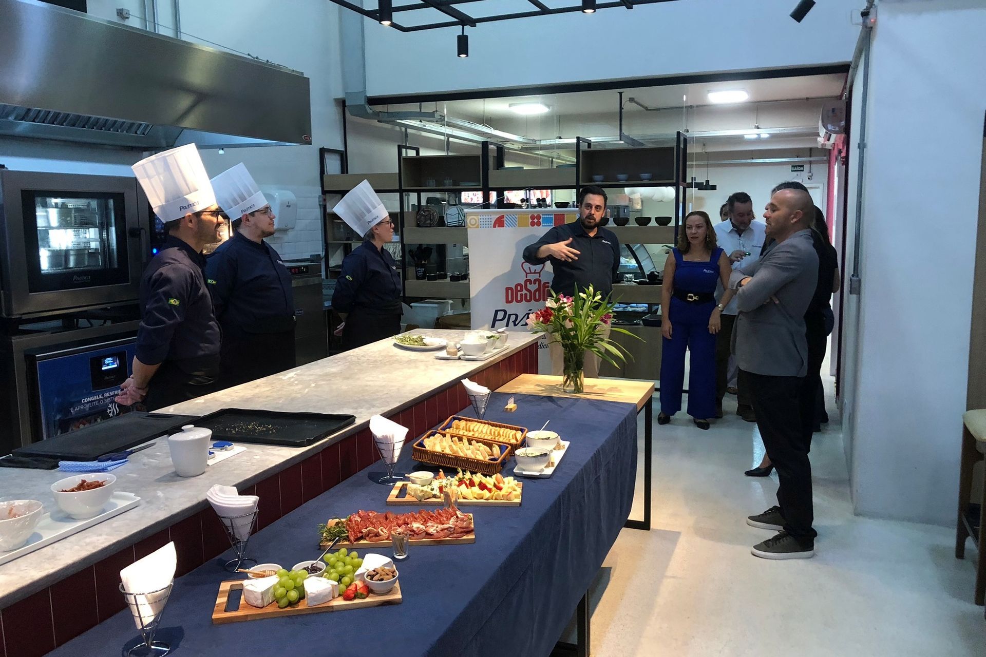 Chefs de chapéu branco e avental ouvem um homem falar em um ponto de distribuição de alimentos.