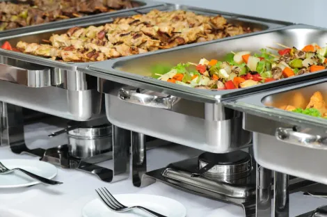 Buffet com bandejas de aço inoxidável contendo pratos quentes e saladas em rechauds, com garfos sobre uma toalha de mesa branca.