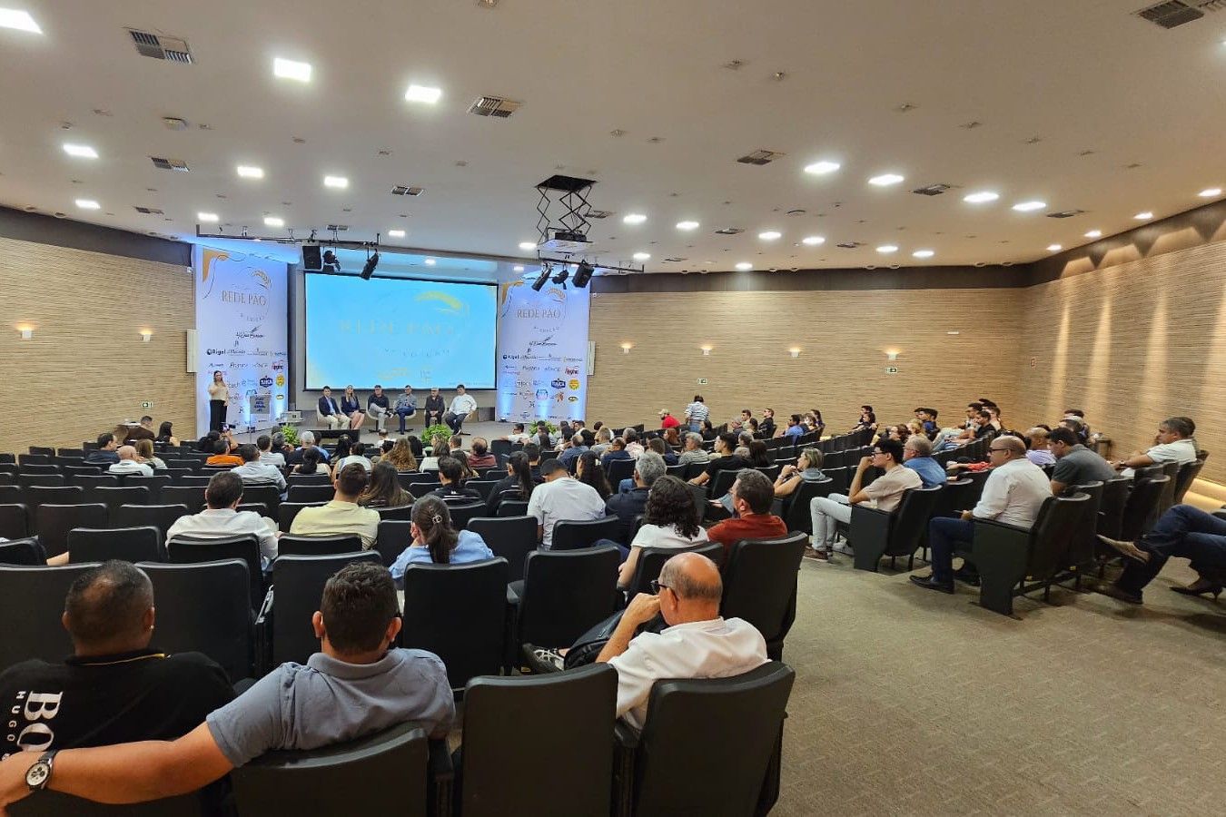 Um grande auditório cheio de pessoas sentadas em cadeiras assistindo a uma apresentação.