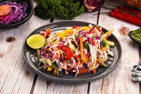 Um prato de salada de repolho colorida com repolho roxo desfiado, pimentões e uma fatia de limão sobre uma mesa de madeira.