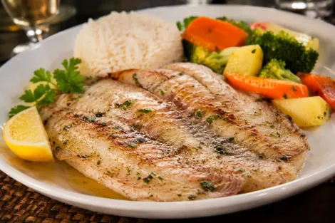 Filé de peixe saint peter grelhado com arroz, fatia de limão e legumes cozidos no vapor em um prato branco.