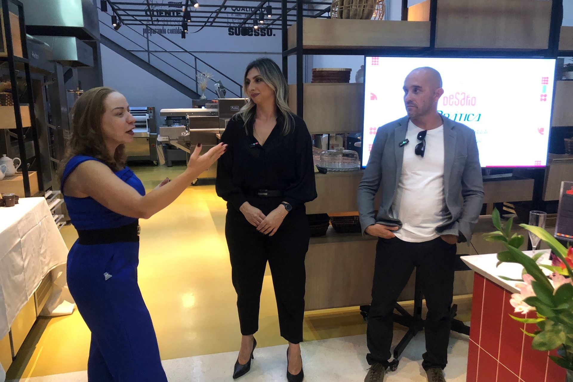 Três pessoas em pé em um espaço interior. Uma mulher gesticula, enquanto outras duas ouvem atentamente. Ao fundo, um painel de apresentação.