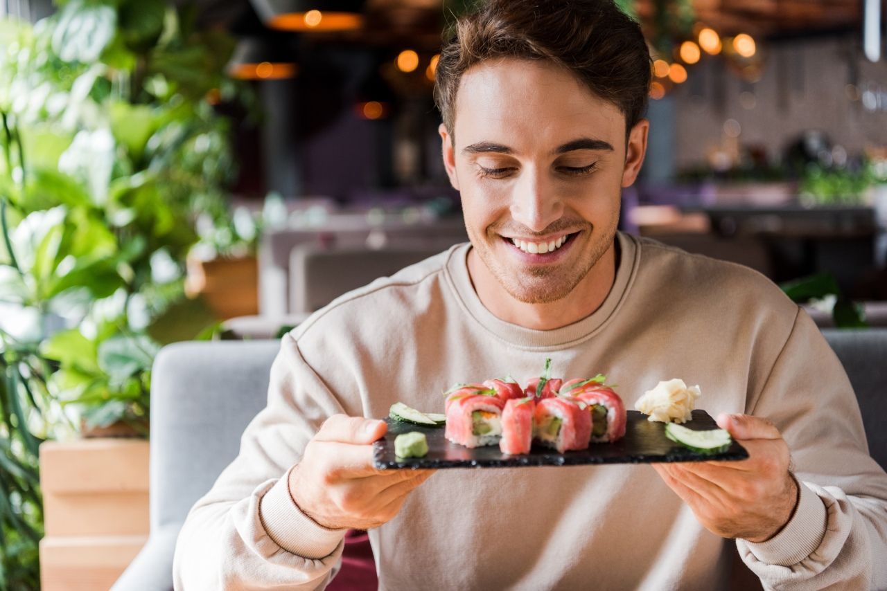 Homem sorridente segurando um prato de sushi em um restaurante iluminado.