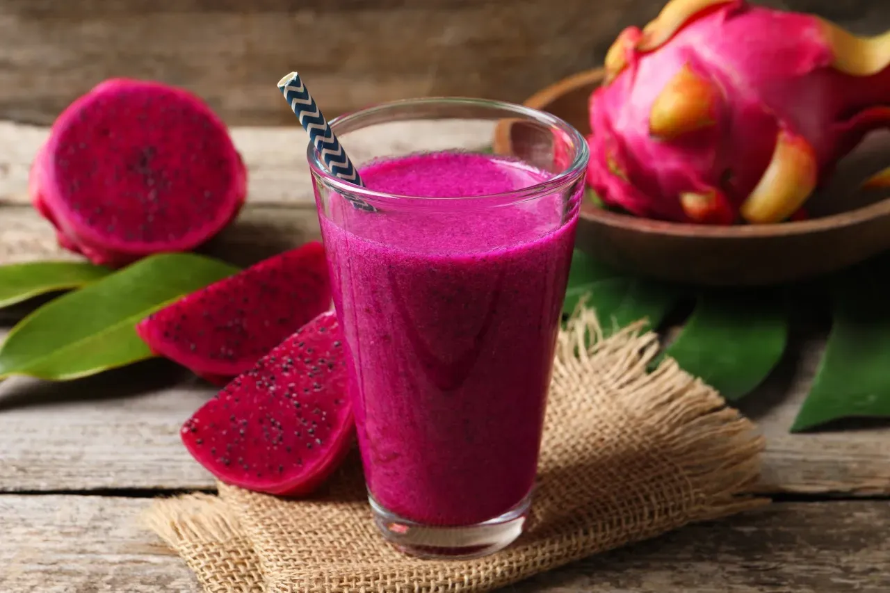 Copo de suco de pitaya rosa vibrante com canudo, pedaços de pitaya e frutas inteiras sobre uma superfície de madeira.