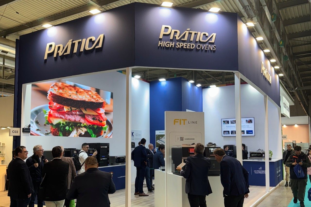 Estande da Pratica em uma feira comercial, exibindo uma grande foto de um hambúrguer; pessoas reunidas dentro e ao redor do estande.