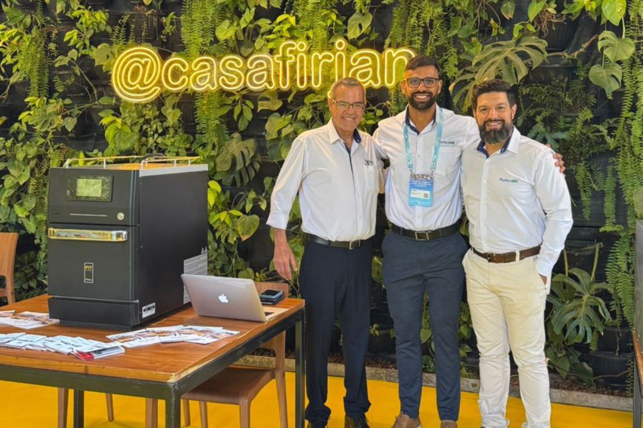 Três homens posam com uma máquina em um evento; um no meio usa um crachá e um laptop está sobre uma mesa.