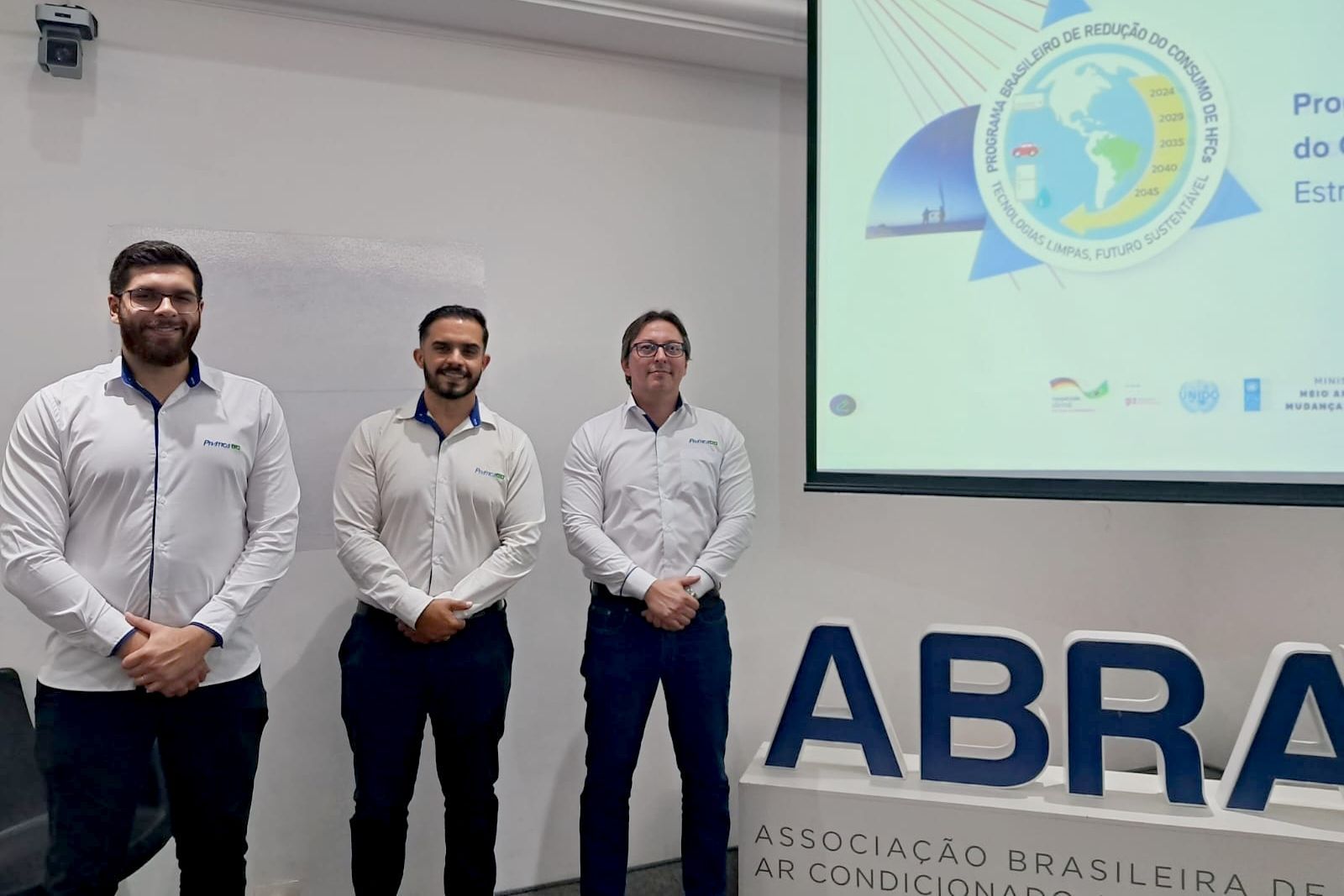 Três homens de camisa branca e calça escura estão em frente a uma tela de projeção com um logotipo e texto, e uma placa que diz 