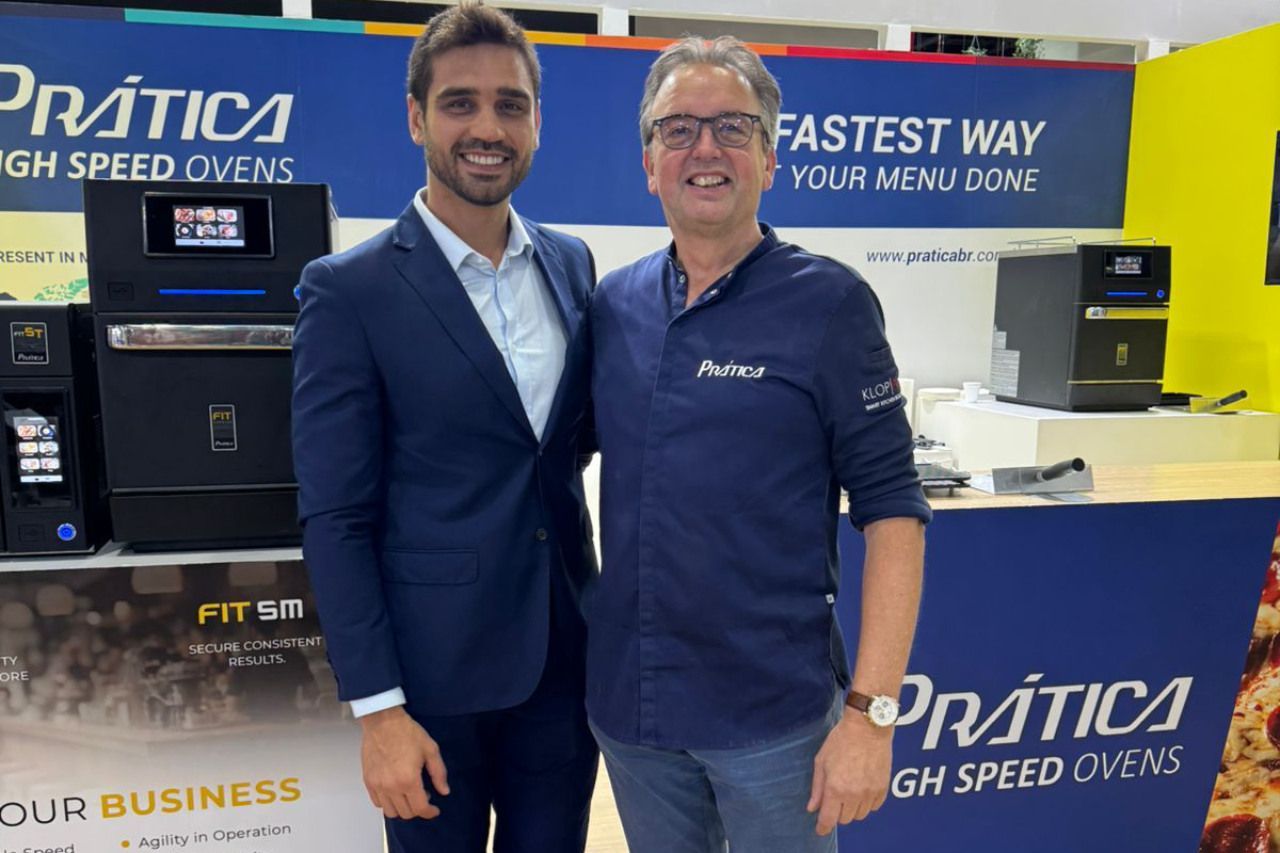 Dois homens posam para uma foto em frente a uma placa que diz prática