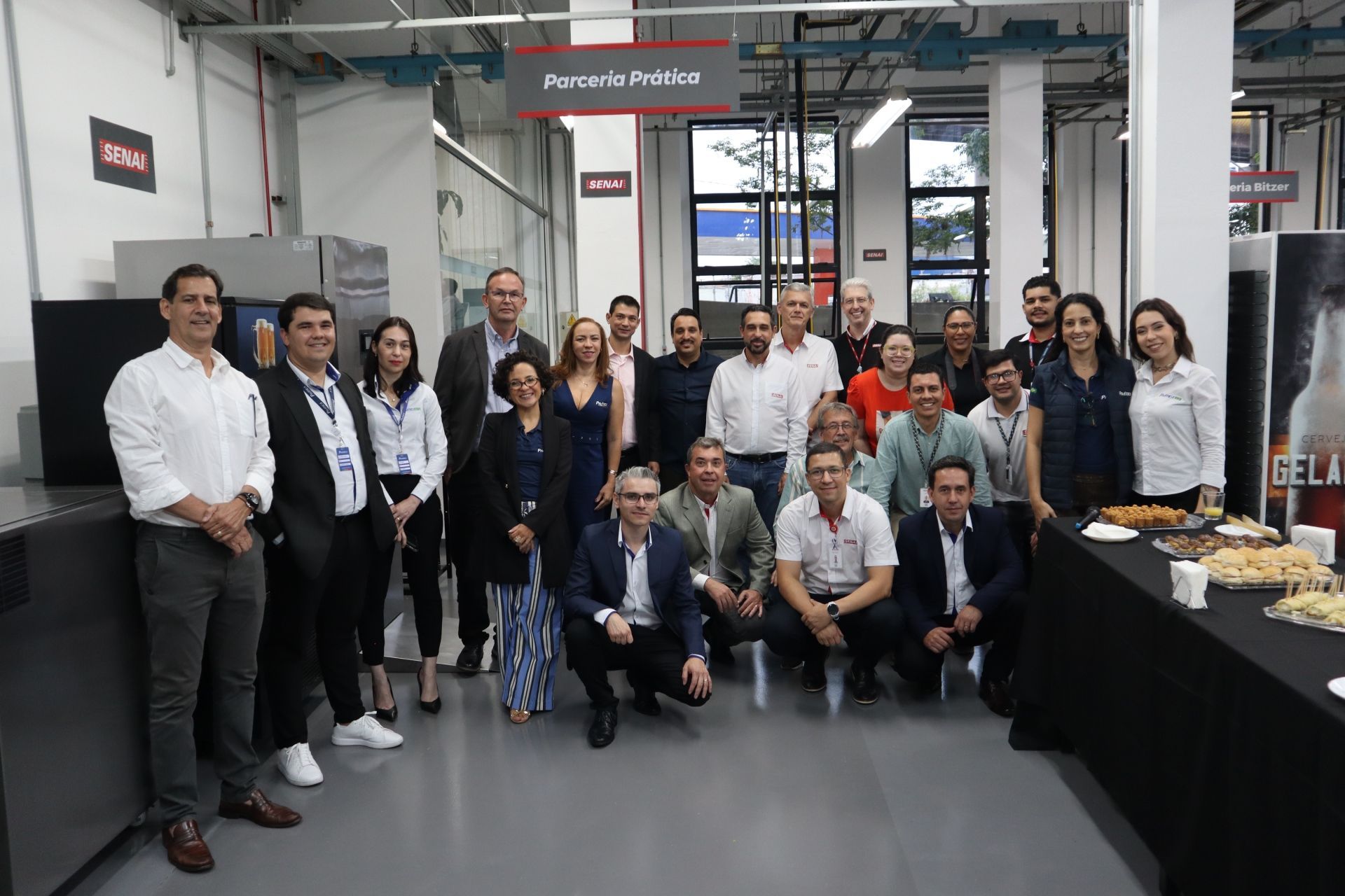 Grupo de pessoas posando para uma foto em um ambiente de workshop. Algumas estão sorrindo, outras vestem trajes formais.