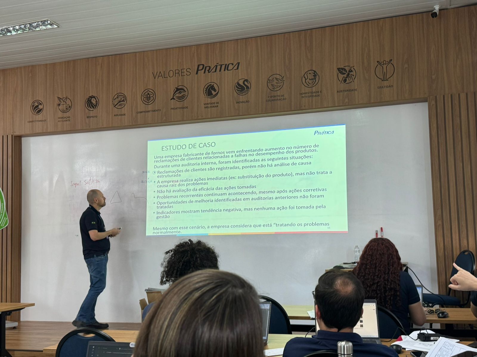 A person stands by a projected screen displaying bulleted text during a presentation in a wood-paneled classroom.