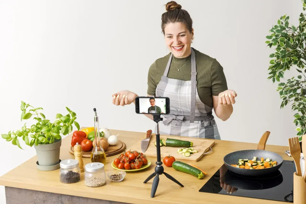 Pessoa cozinhando em uma bancada, sorrindo ao lado de ingredientes frescos e um celular em um tripé, com uma panela no fogão.