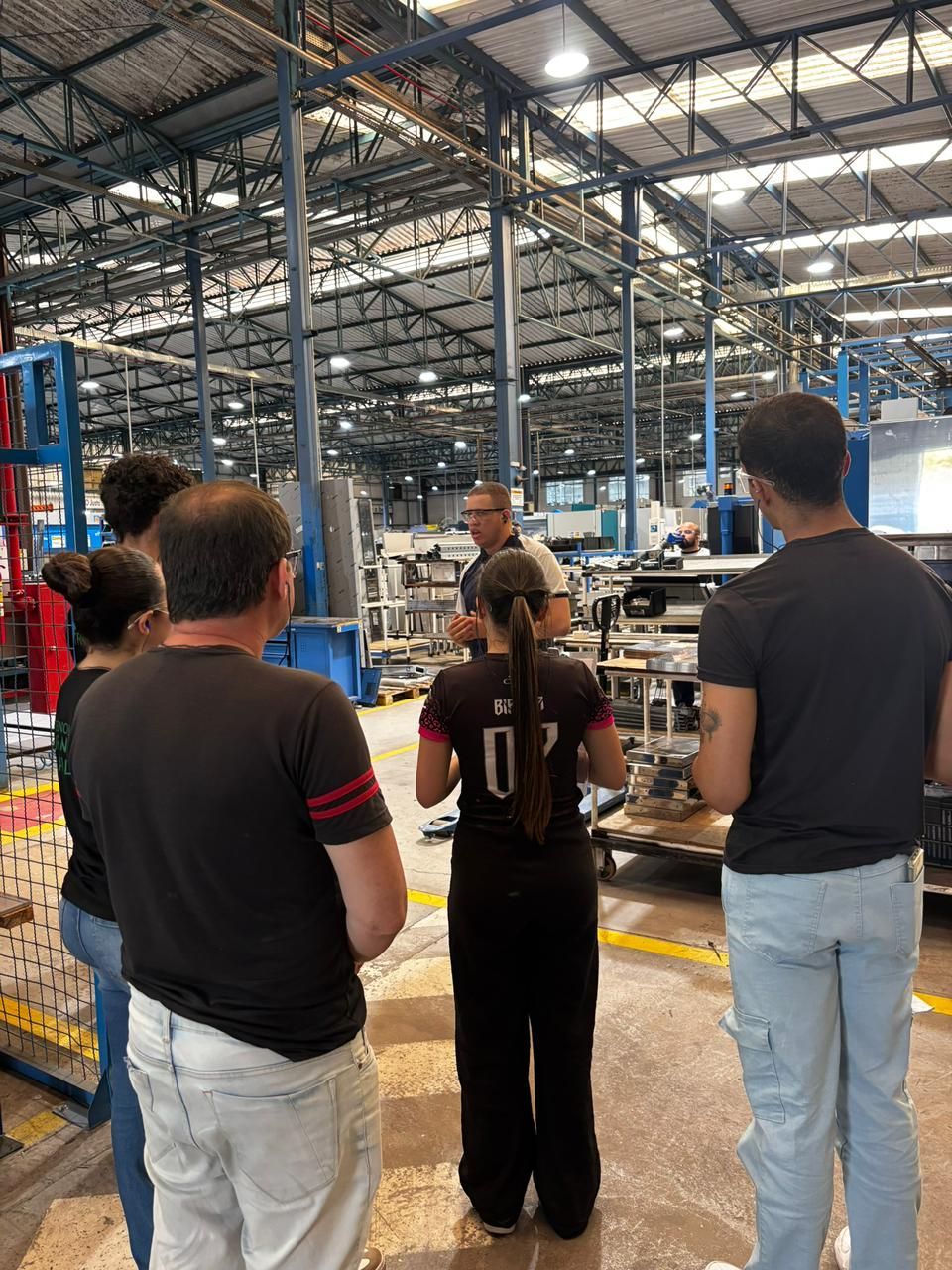 Um grupo de pessoas ouve um homem em um ambiente fabril, perto de equipamentos e prateleiras de metal.