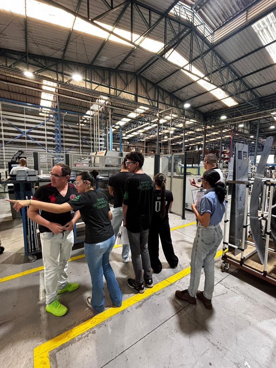 Pessoas em uma fábrica, observando as máquinas. Um homem aponta para a esquerda. Alguns vestem camisas pretas, outros calças jeans. Há uma linha amarela no chão.